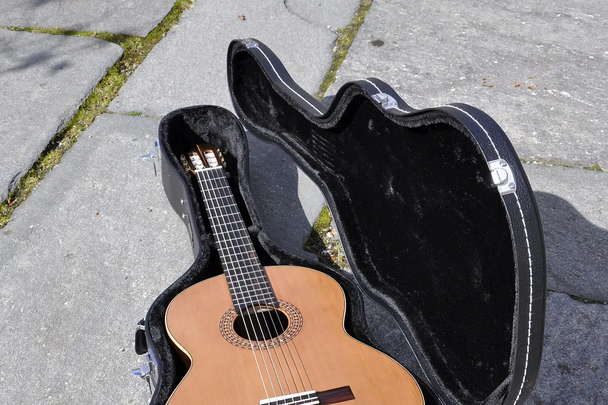 A classical guitar with a pale spruce top and nylon strings lying in an open black case on paving stones